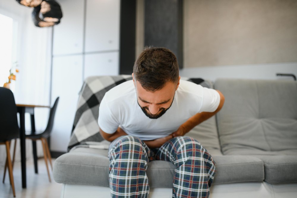 גבר יושב על ספה ואוחז בבטן התחתונה בתחושת כאב ואי נוחות, המחשה של כאבי בטן ועצירות בסביבה ביתית. גבר יושב על ספה ואוחז בבטן התחתונה בתחושת כאב ואי נוחות, המחשה של כאבי בטן ועצירות בסביבה ביתית.